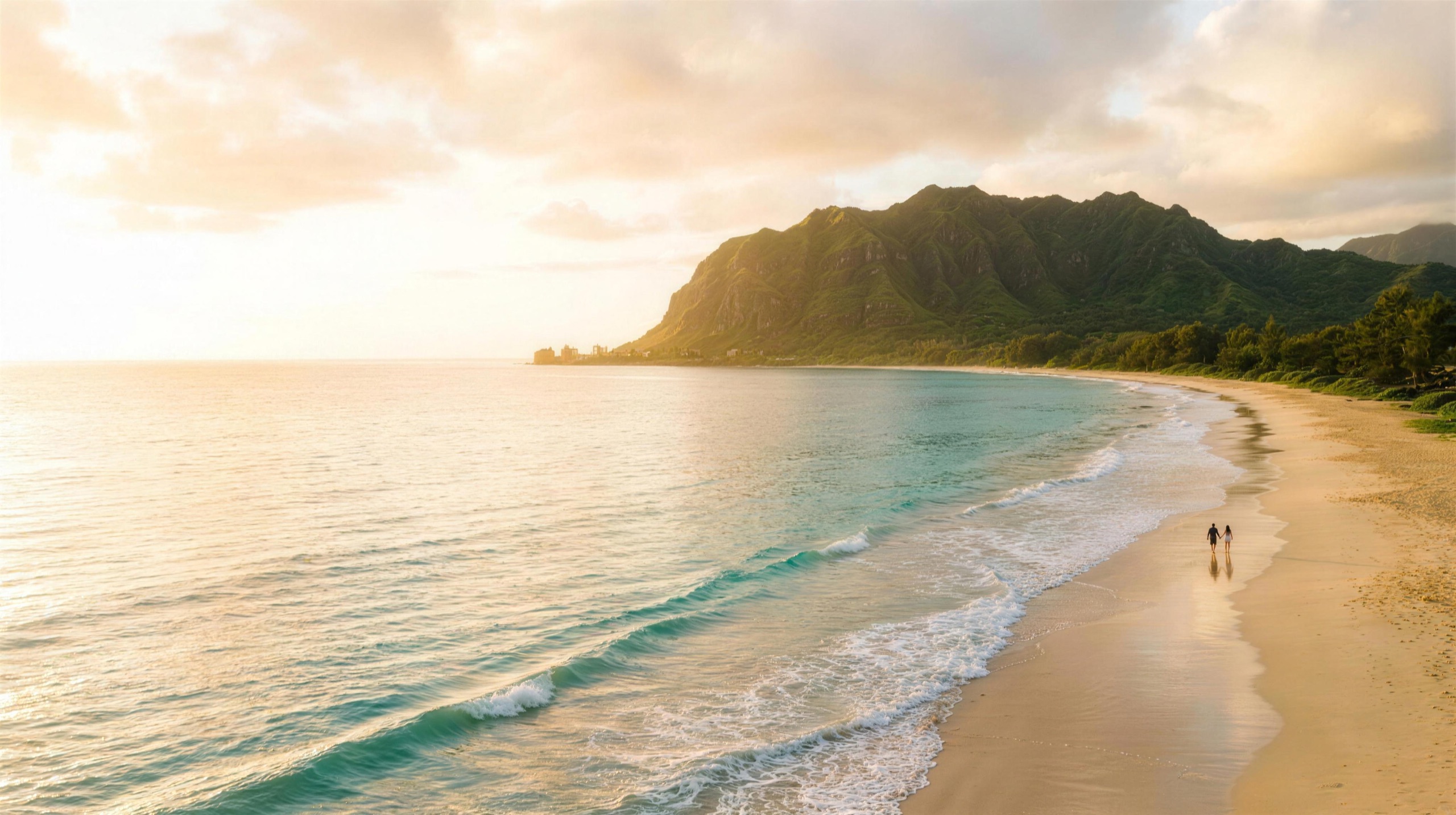 Golden-hour aerial of an Oʻahu beach — turquoise water, lush windward ridge, and a couple walking hand-in-hand along the shore